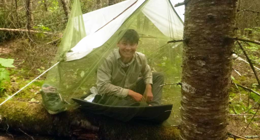 A student sits and smiles from under their tarp shelter in a wooded area. The shelter is covered in a mosquito net. 